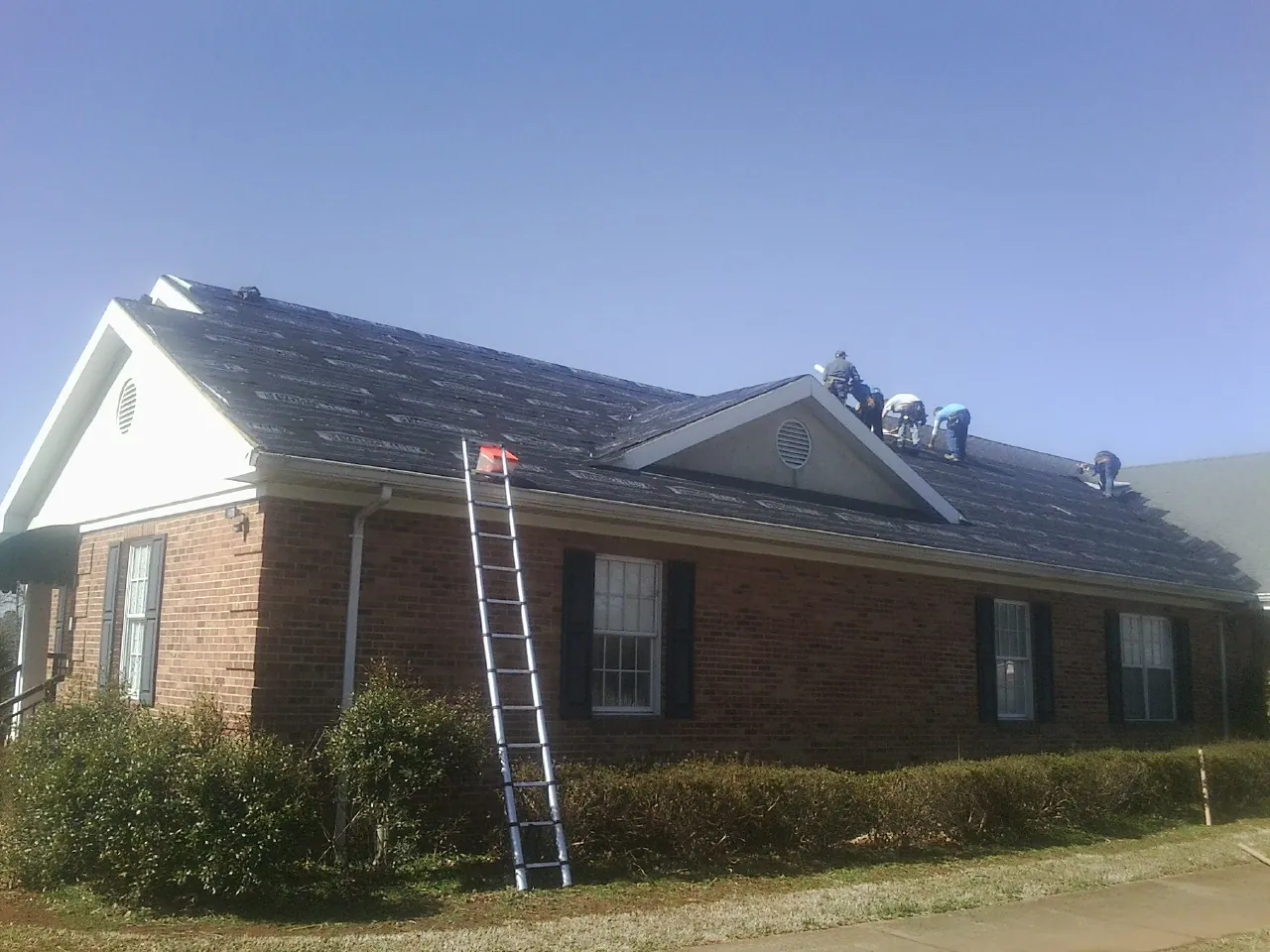 Crew tearing shingles off a brick ranch roof with a ladder set — Emergency Roof Repair work by our crew in Brimfield